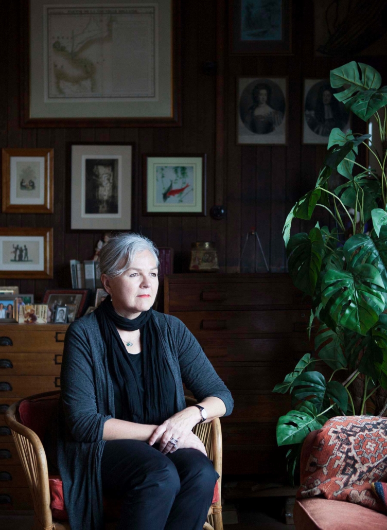 woman with grey hair sits on a chair, looking outside with artwork and plants behind her.