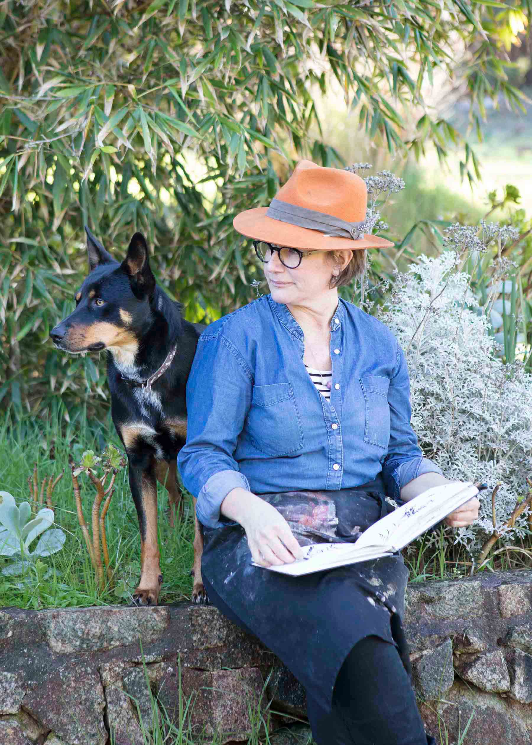 woman with orange hat sits to draw in an open book with her dog beside her